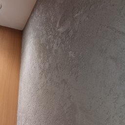 Jakub Jabłoński - Szara ściana z dekoracyjną fakturą, widoczny narożnik z drewnianą boazerią i biały sufit z lampą.