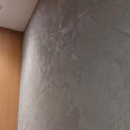 Szara ściana z dekoracyjną fakturą, widoczny narożnik z drewnianą boazerią i biały sufit z lampą.