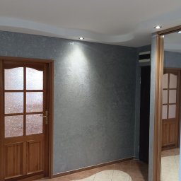 Jakub Jabłoński - Ściana w szarym kolorze z efektem dekoracyjnym, oświetlona punktowym światłem, drewniane drzwi z szybą w kratkę, podłoga z płytek w odcieniach beżu i brązu, odbicie w lustrze przesuwnych drzwi szafy.