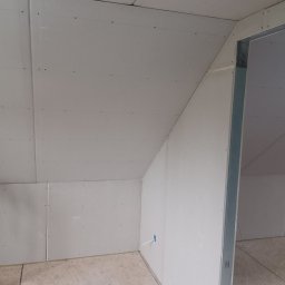Jakub Jabłoński - Poddasze z zamontowaną płytą kartonowo-gipsową, widoczne łączenia płyt i otwór na instalację elektryczną, surowy stan wykończenia, metalowa ościeżnica drzwiowa.