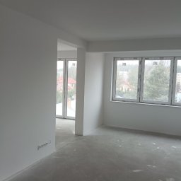 Jakub Jabłoński - Wnętrze w stanie deweloperskim po wykonaniu gładzi i malowania ścian na biało, widoczne okna z widokiem na zabudowę jednorodzinną i wielorodzinną, surowa betonowa podłoga z resztkami gipsu.