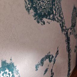 Jakub Jabłoński - Dekoracyjna ściana z efektem przetarć, z motywami mandali w odcieniach turkusu na teksturowanym, jasnym tle. Artystyczne wykończenie wnętrza, widok z perspektywy.