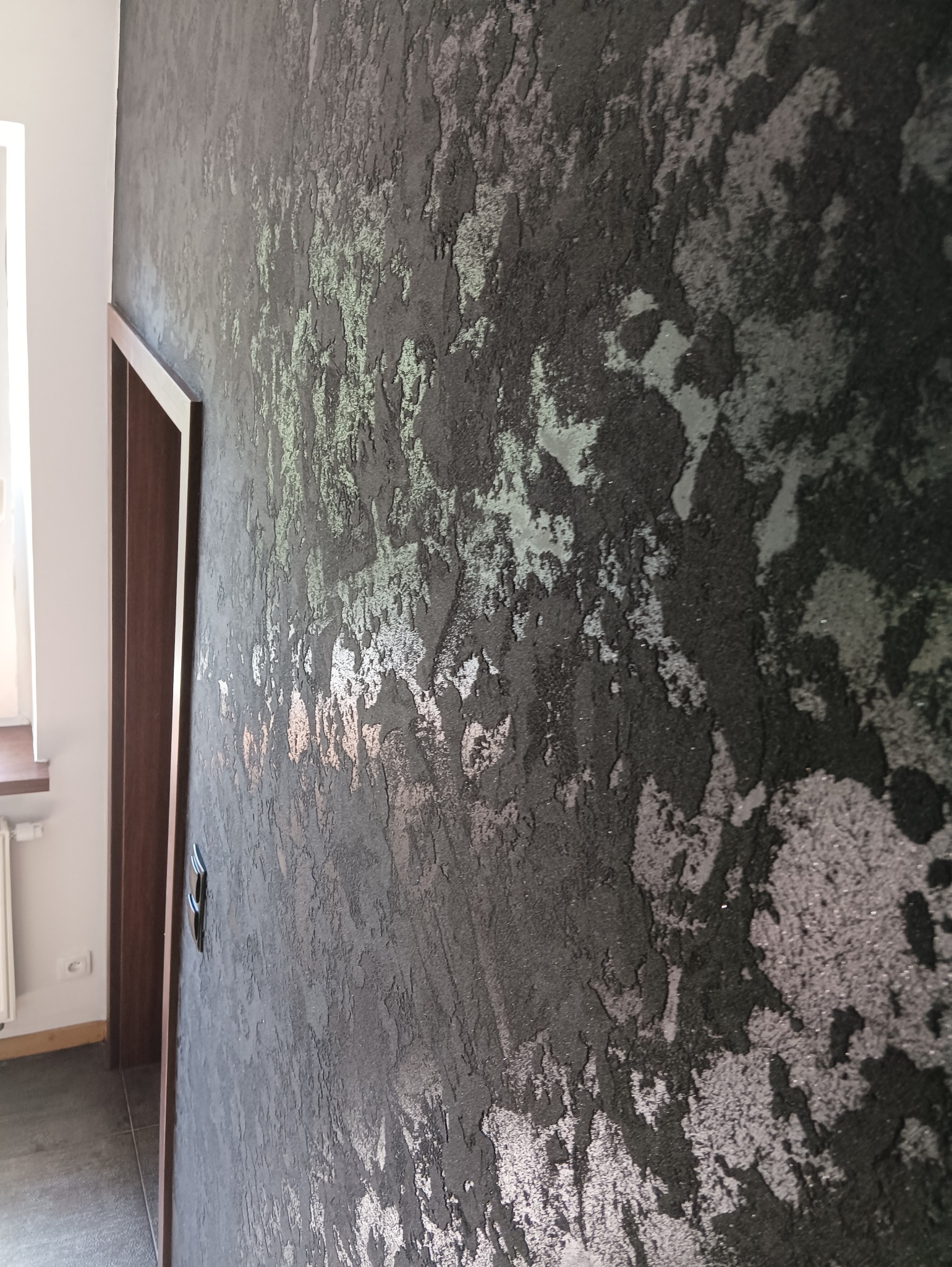 Dekoracyjna ściana z efektem strukturalnym w odcieniach szarości i srebra, widoczna obok futryny drzwiowej i fragmentu okna.