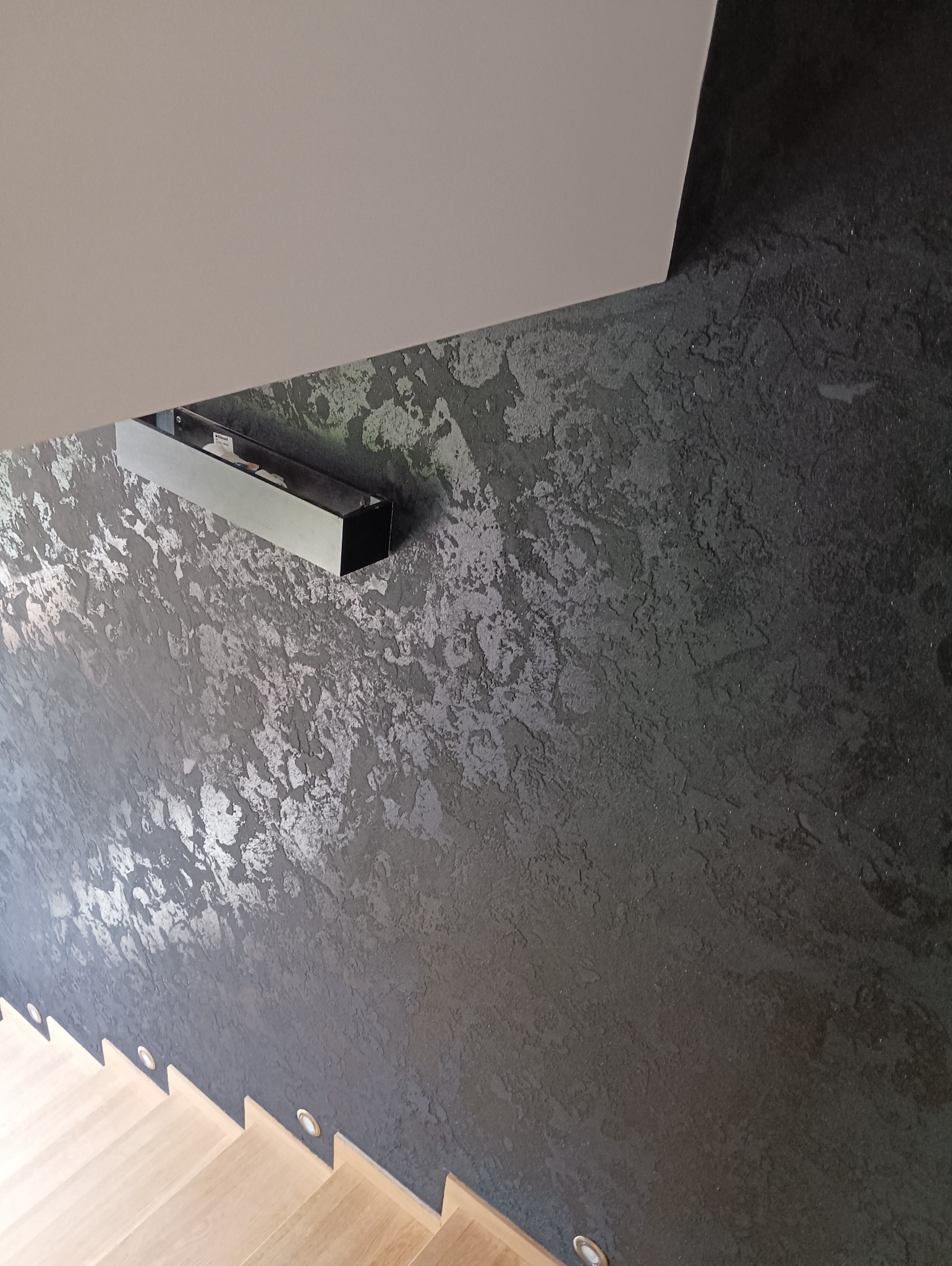 Ściana wykończona dekoracyjną, ciemną strukturą z srebrnymi refleksami, oświetlona listwą LED i punktowymi lampkami schodowymi, widok z góry na drewniane schody.