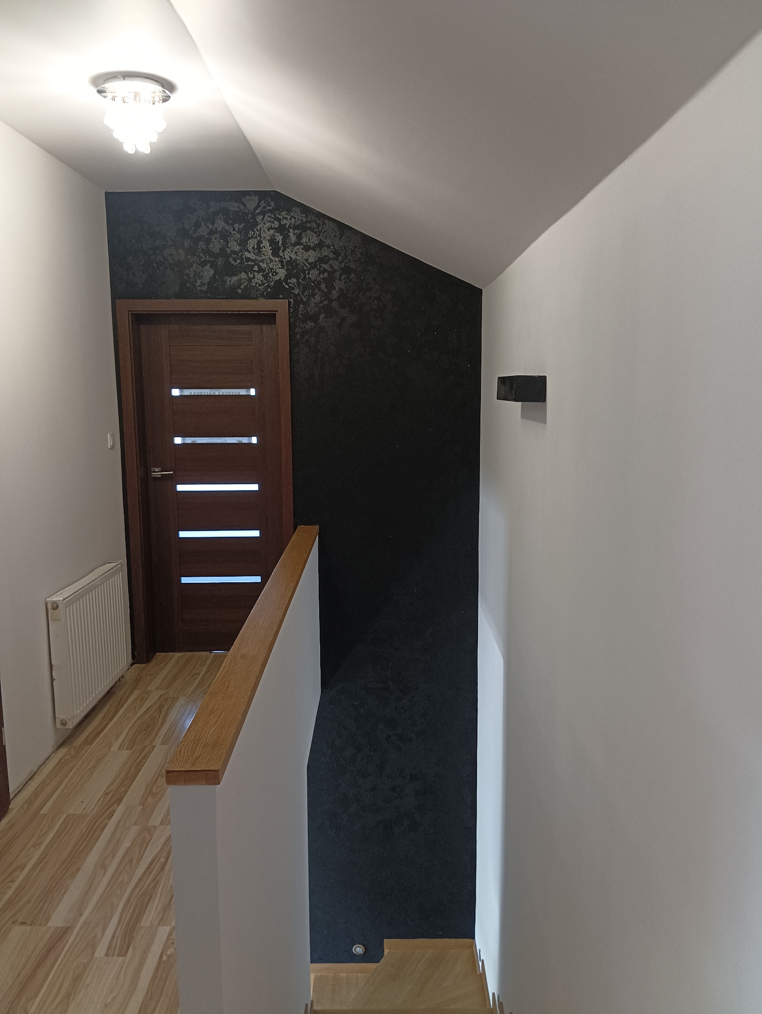 Korytarz w domu z wykończonymi ścianami: gładka, biała ściana kontrastuje z ciemną ścianą pokrytą dekoracyjną strukturą, widoczna drewniana balustrada schodów, drzwi oraz fragment grzejnika.
