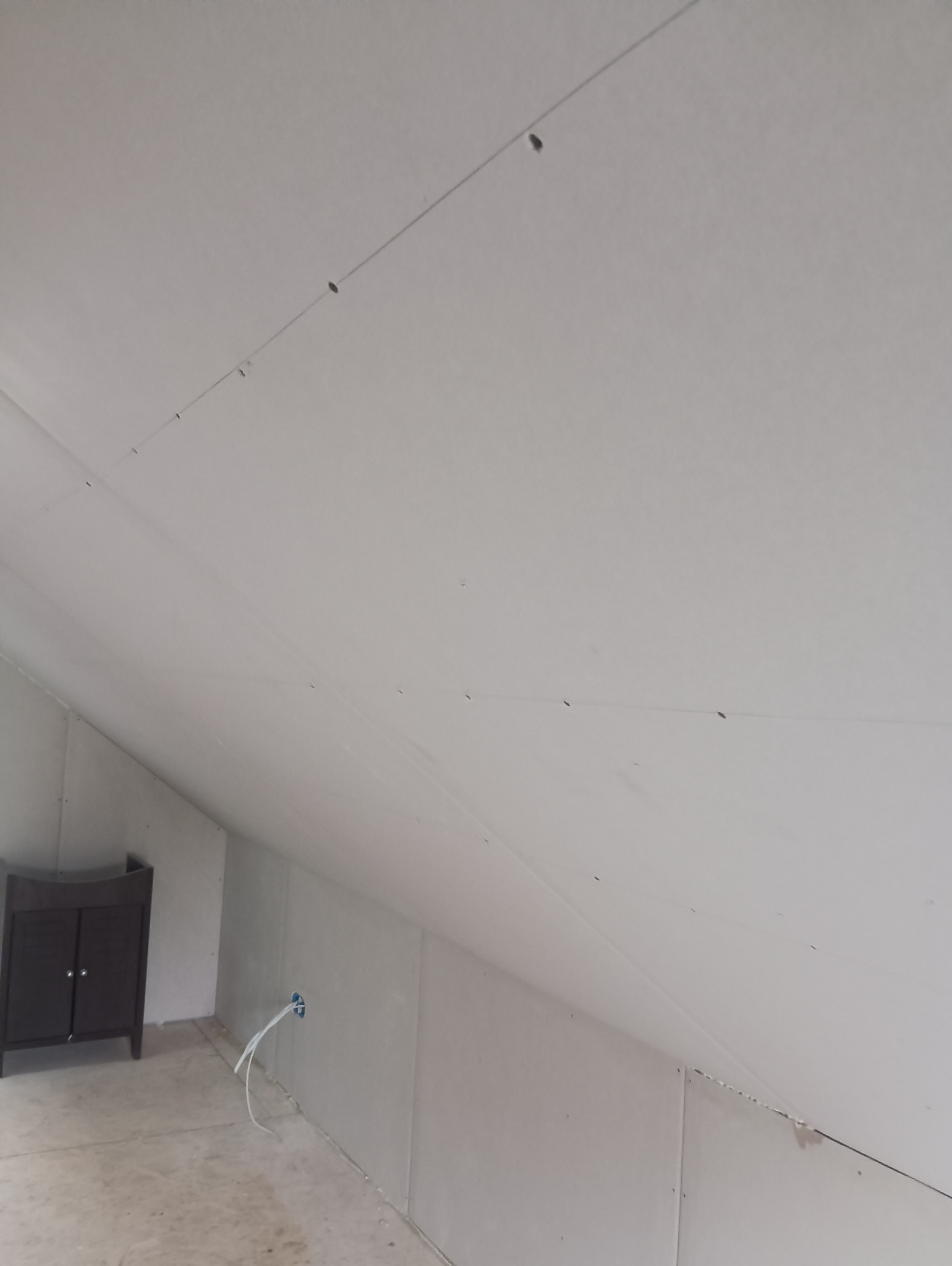 Poddasze z zamontowaną płytą kartonowo-gipsową na skosach, widoczne ślady po wkrętach, wystające kable ze ściany i szafka w rogu.