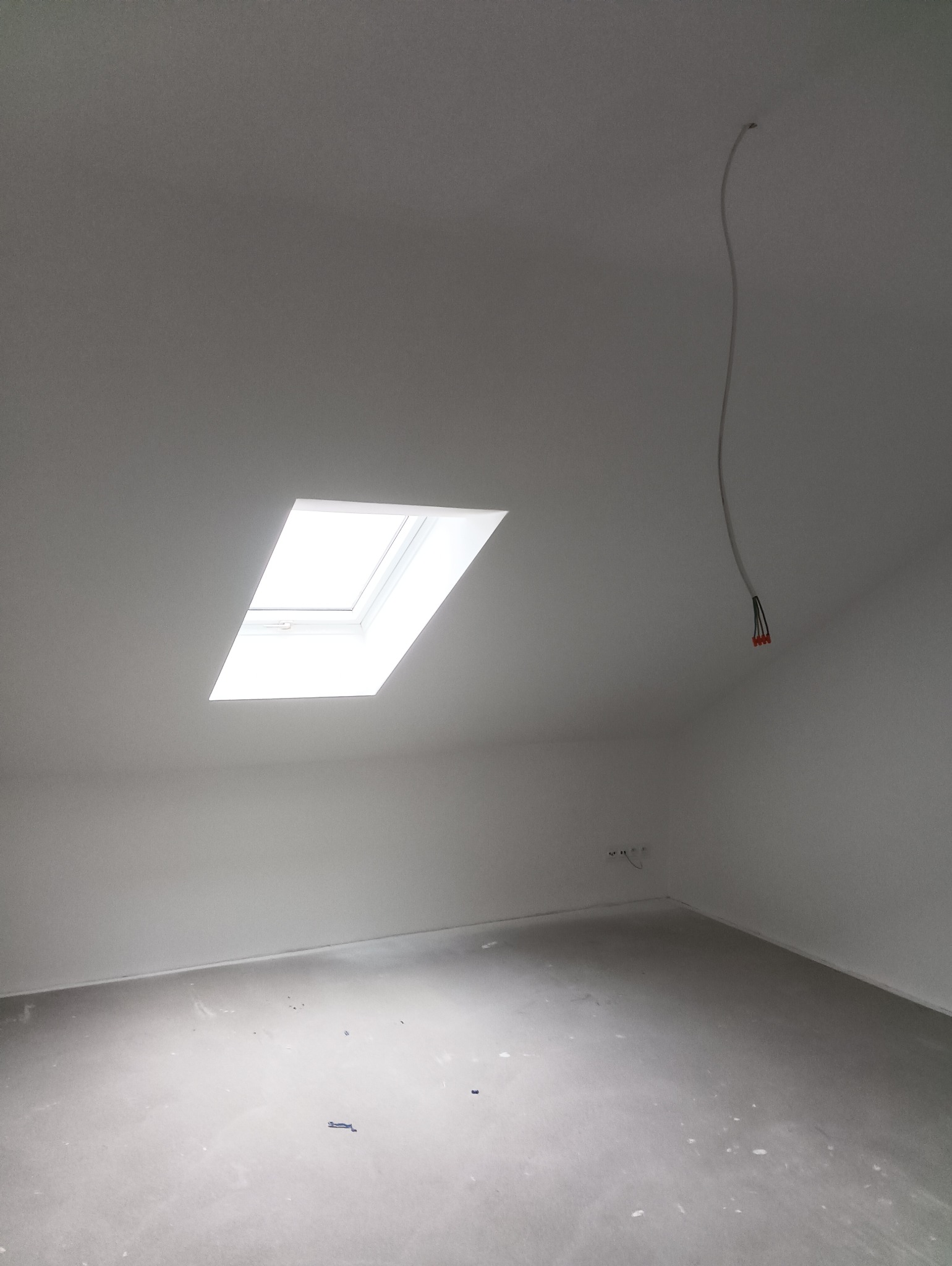 Poddasze po świeżym pomalowaniu na biało, widoczne okno dachowe i wystające kable ze ściany, podłoga w trakcie przygotowania.