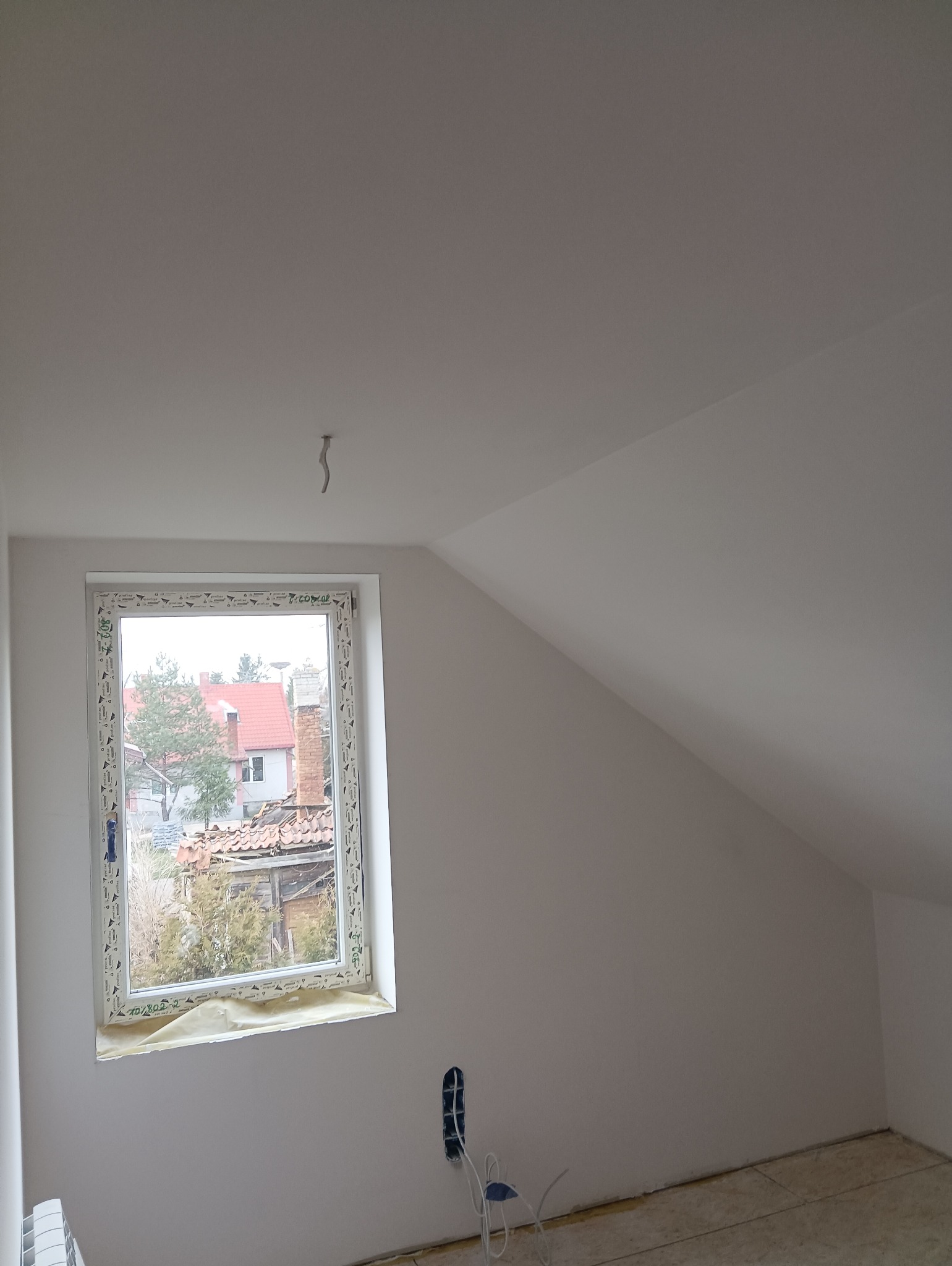 Poddasze po pracach wykończeniowych: świeżo pomalowane ściany w odcieniach bieli, okno z widokiem na zabudowę jednorodzinną, zabezpieczone folią parapety, widoczne gniazdko elektryczne...