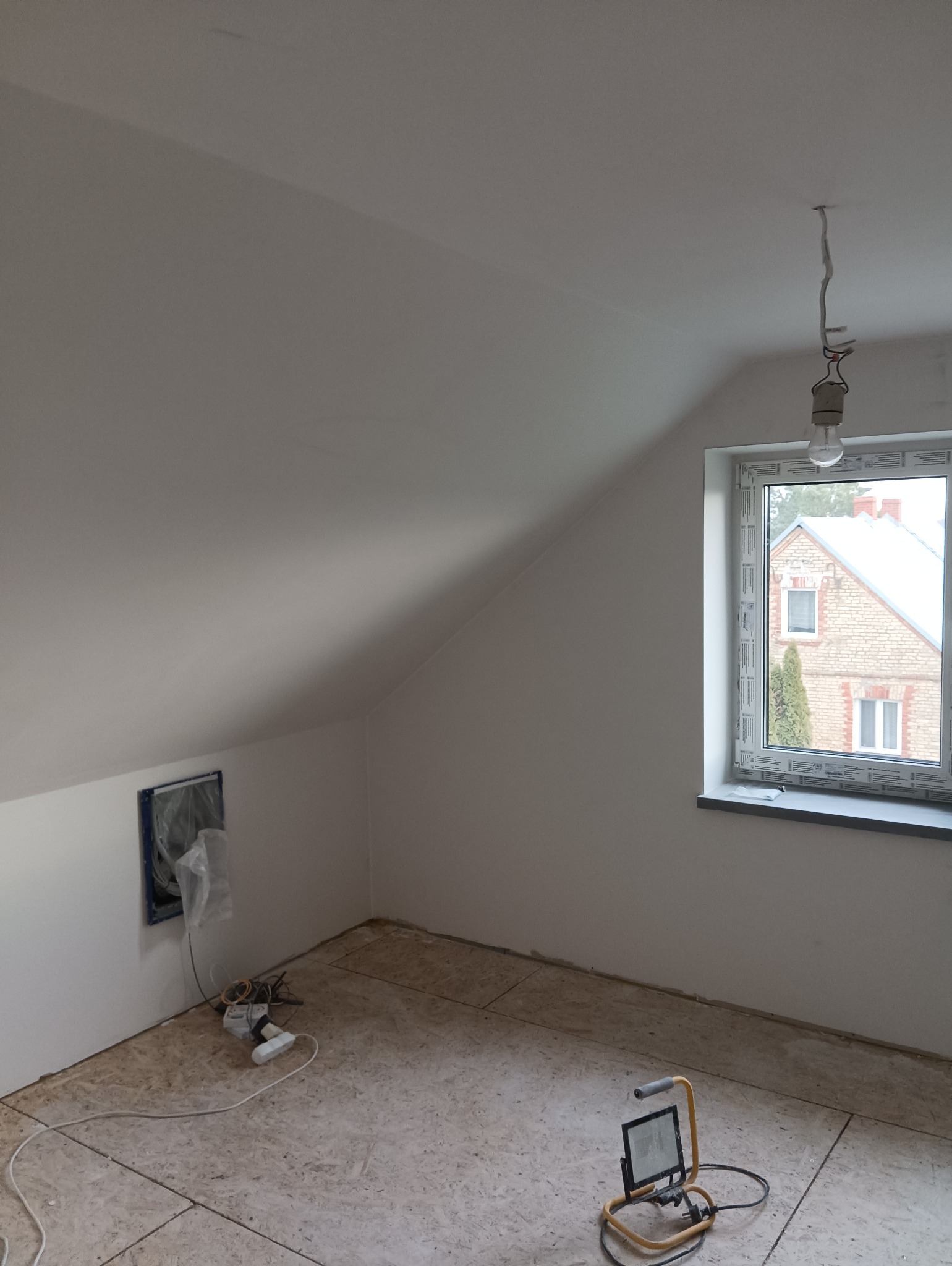 Poddasze po nałożeniu gładzi, z oknem PCV, widoczną instalacją elektryczną, lampą roboczą i podłogą z płyt OSB przygotowaną pod dalsze prace wykończeniowe.