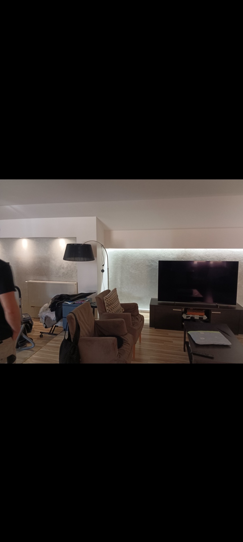 Salon z szarą ścianą, oświetleniem LED, brązowymi fotelami, telewizorem na szafce RTV i podłogą z paneli. W tle widoczny grzejnik i rzeczy osobiste, sugerujące trwające prace.