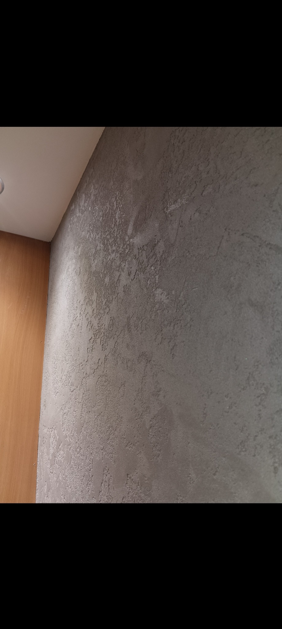 Szara ściana z dekoracyjną fakturą, widoczny narożnik z drewnianą boazerią i biały sufit z lampą.