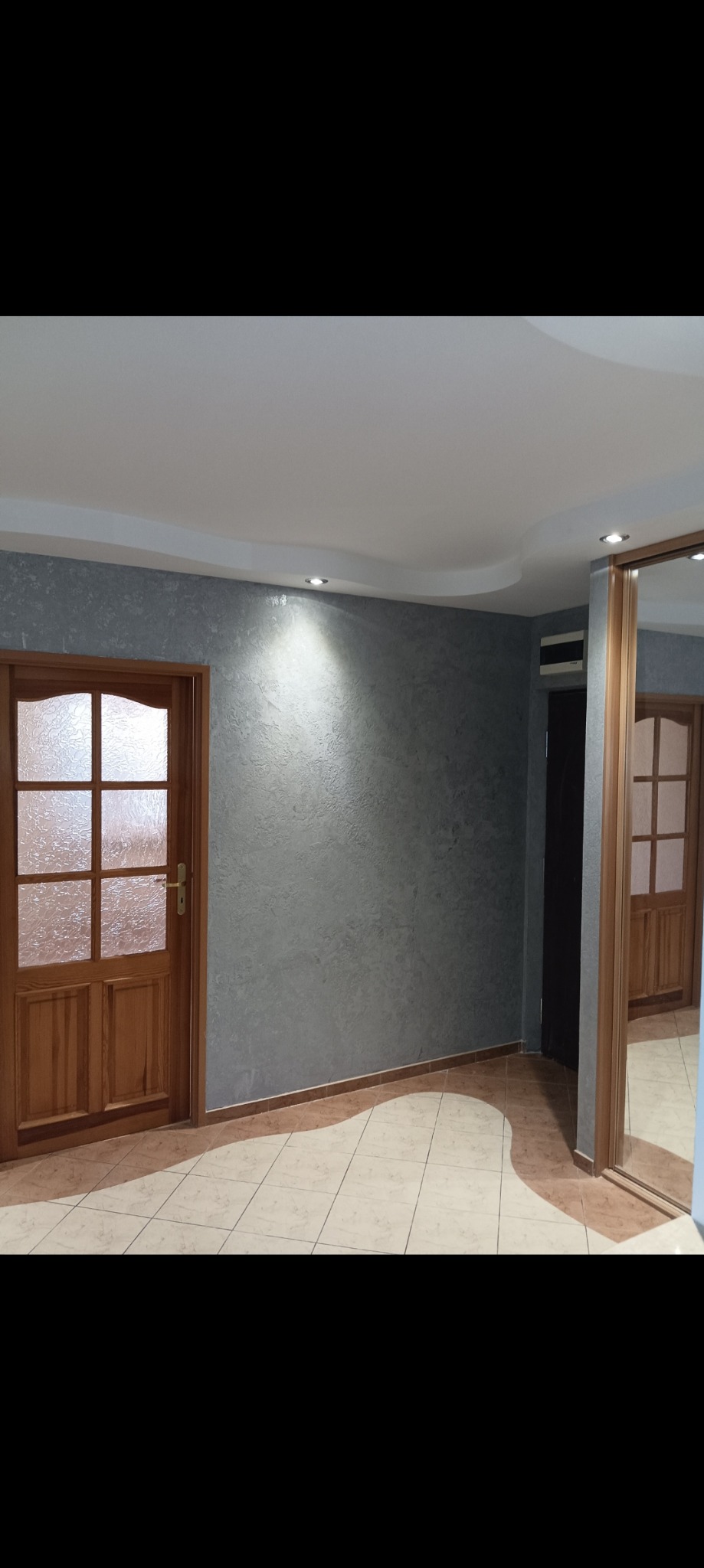 Ściana w szarym kolorze z efektem dekoracyjnym, oświetlona punktowym światłem, drewniane drzwi z szybą w kratkę, podłoga z płytek w odcieniach beżu i brązu, odbicie w lustrze przesuwnych drzwi szafy.