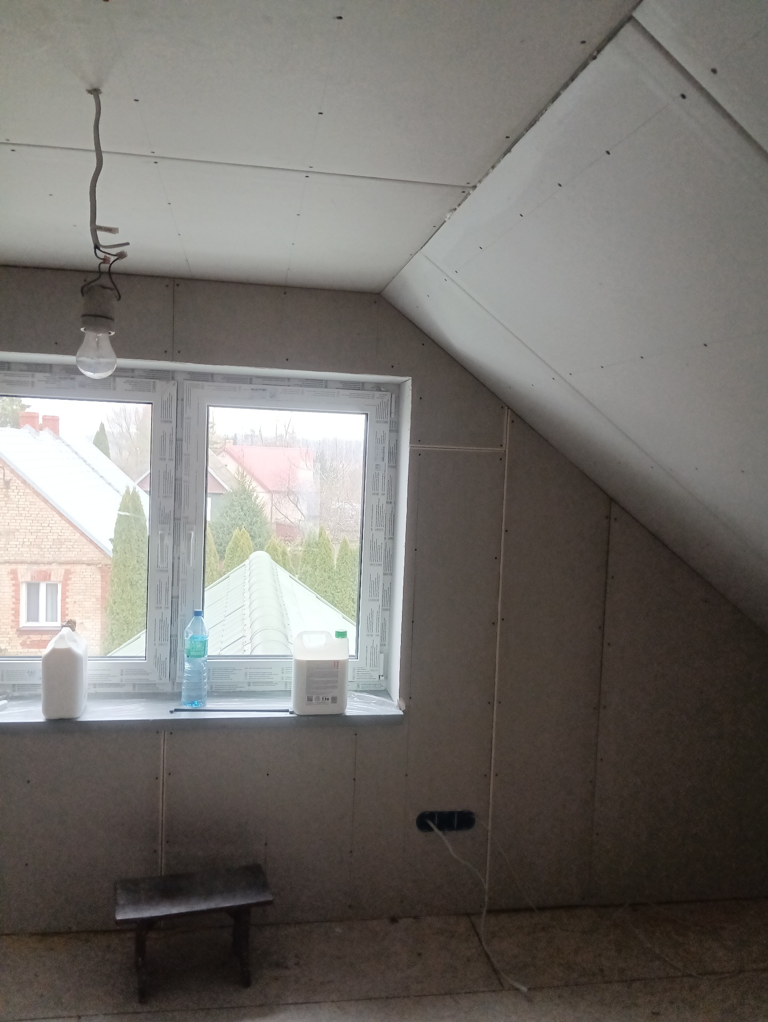 Poddasze w trakcie prac wykończeniowych: ściany i skosy obłożone płytami kartonowo-gipsowymi, na parapecie okna stoją pojemniki z materiałami budowlanymi, a z sufitu zwisa żarówka na kablu.