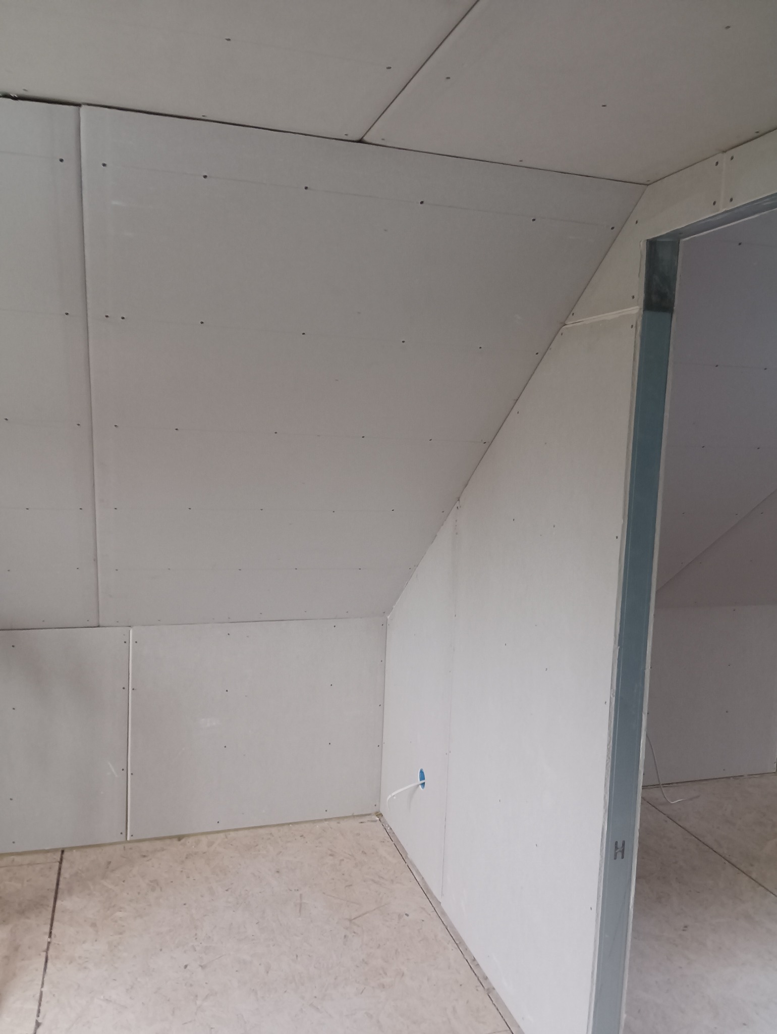 Poddasze z zamontowaną płytą kartonowo-gipsową, widoczne łączenia płyt i otwór na instalację elektryczną, surowy stan wykończenia, metalowa ościeżnica drzwiowa.