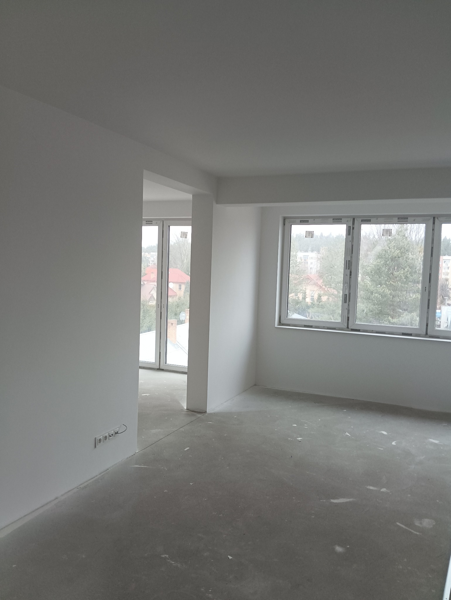 Wnętrze w stanie deweloperskim po wykonaniu gładzi i malowania ścian na biało, widoczne okna z widokiem na zabudowę jednorodzinną i wielorodzinną, surowa betonowa podłoga z resztkami gipsu.