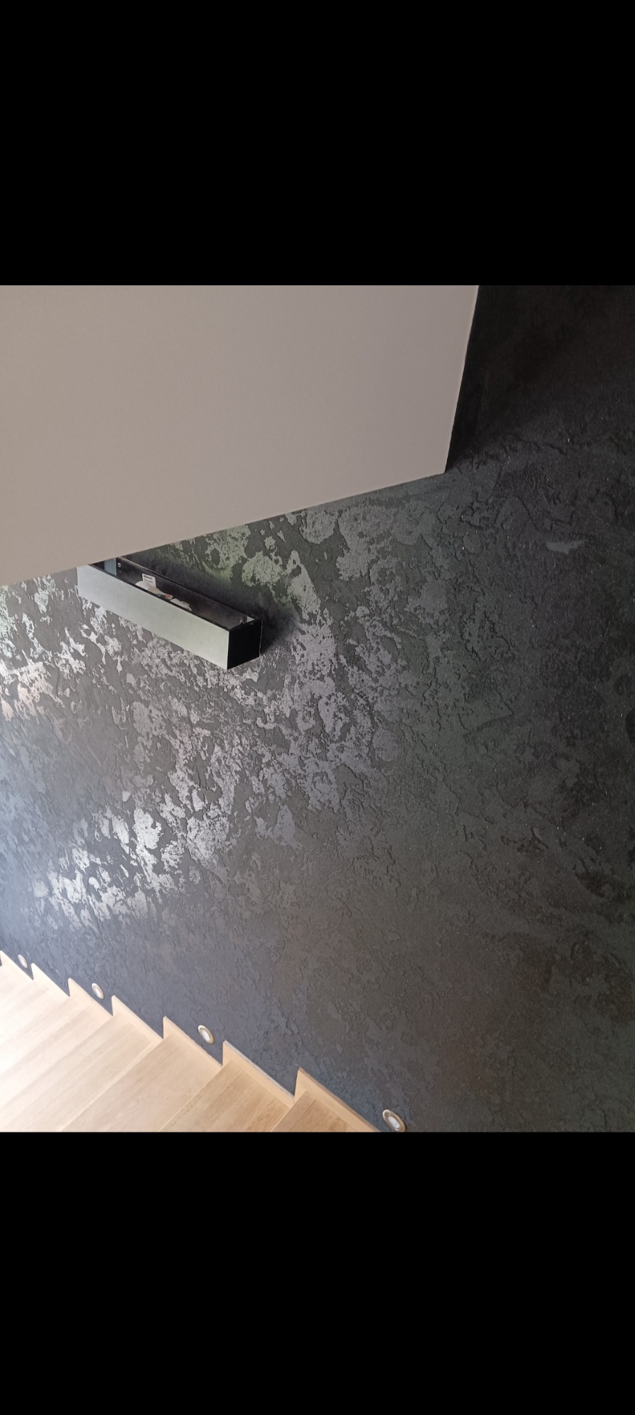 Ściana w ciemnym odcieniu z dekoracyjną fakturą, oświetlona lampą ścienną, obok schody z jasnego drewna z punktowym oświetleniem.