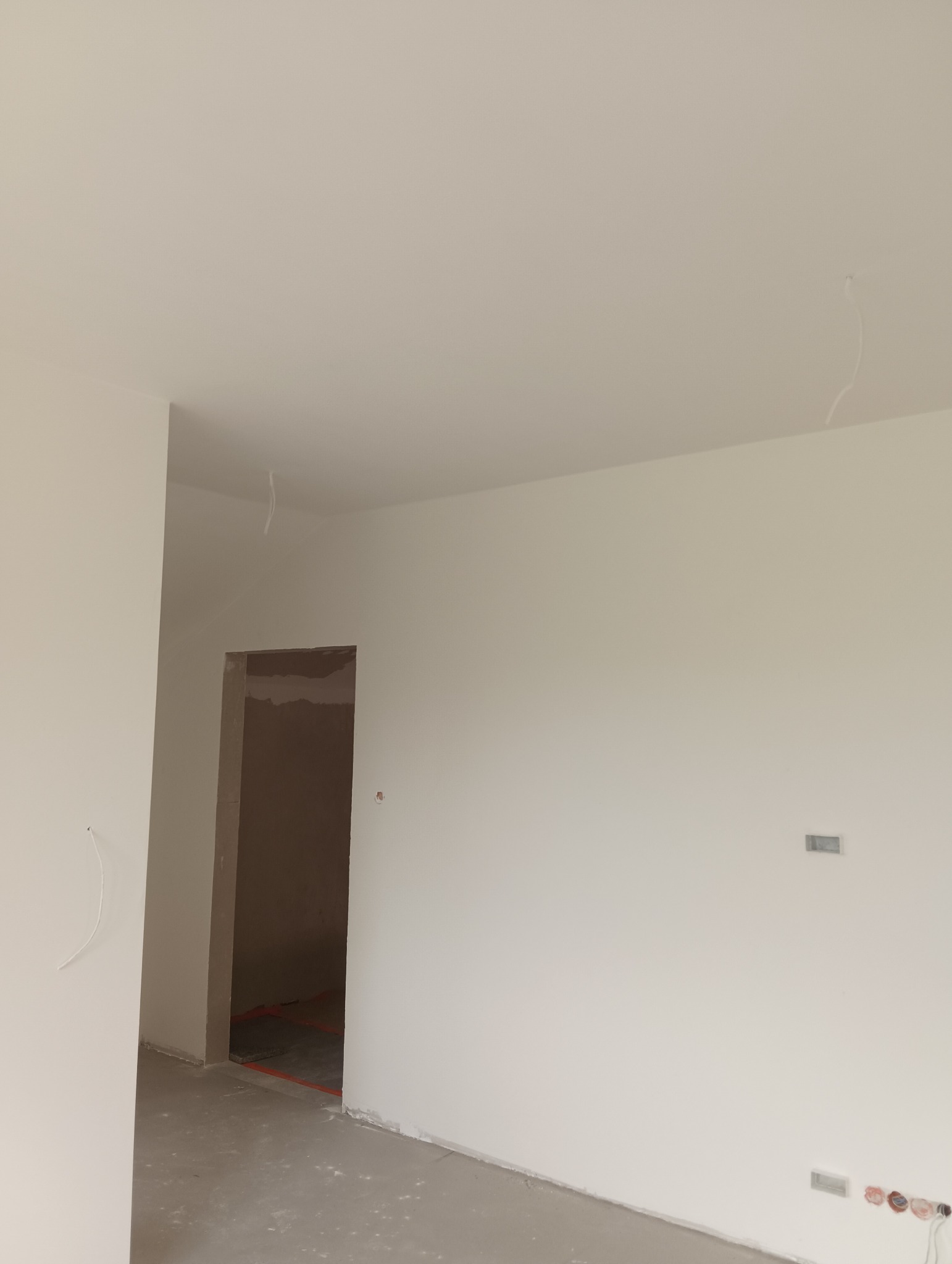 Jasne, świeżo pomalowane wnętrze z widocznymi gniazdkami elektrycznymi i wystającymi kablami. W tle surowy otwór drzwiowy z widoczną instalacją podłogową.