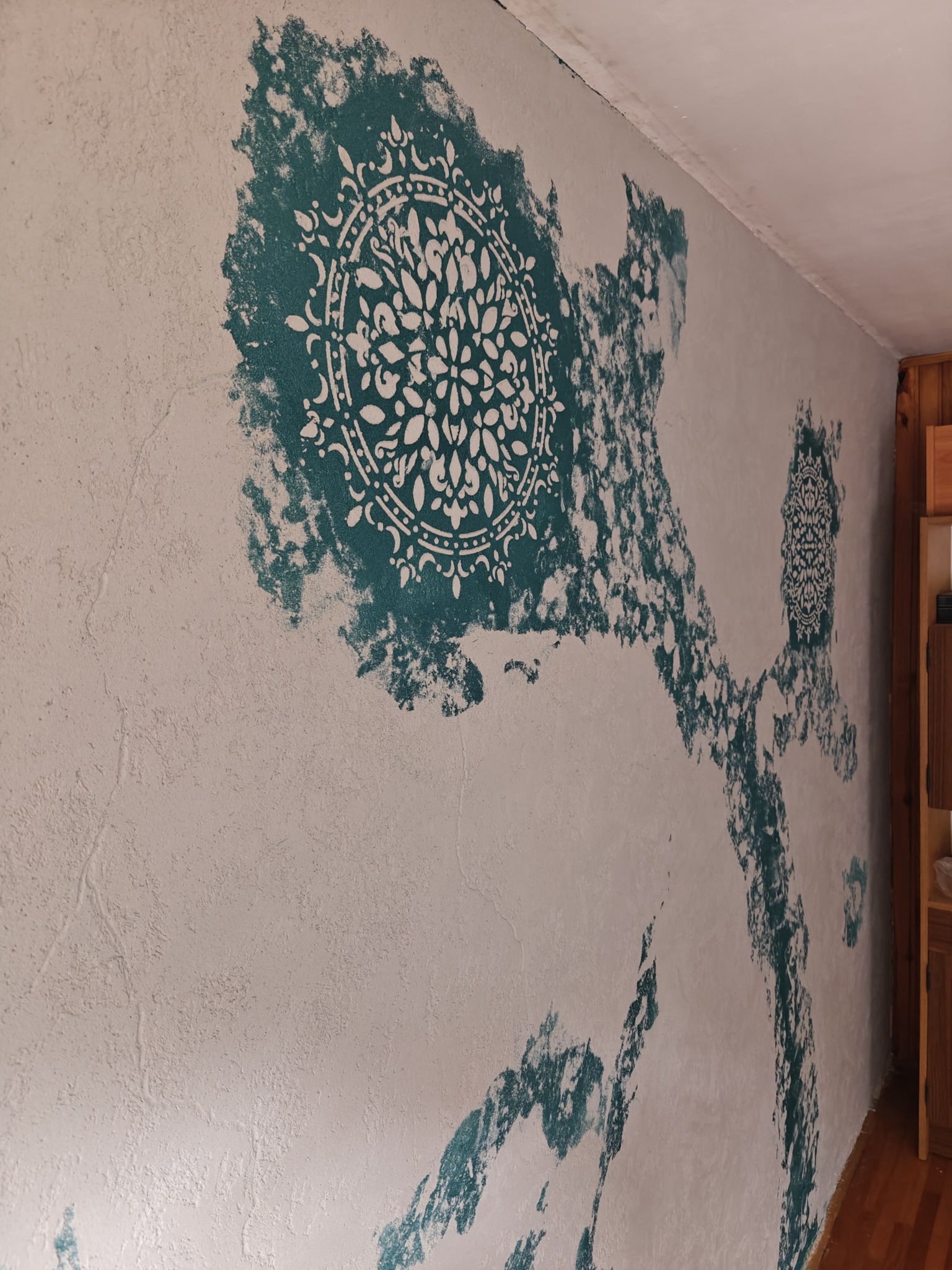 Ściana z dekoracyjną, turkusową aplikacją w stylu mandali, wykonana na nierównej, jasnej powierzchni. Widoczne niedoskonałości i unikalna faktura tynku.