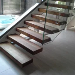 Nowoczesne schody z drewnianymi stopniami i szklaną balustradą, widoczne z boku, z jacuzzi w tle.