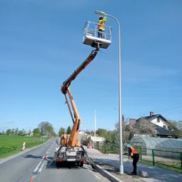 Pracownicy w pomarańczowych kamizelkach montują lampę uliczną za pomocą podnośnika koszowego na tle błękitnego nieba i wiejskiego krajobrazu.