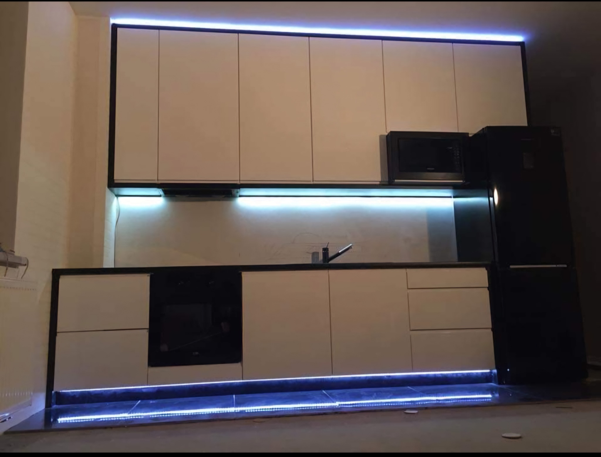 Nowoczesna kuchnia z białymi frontami szafek, czarnymi obramowaniami i oświetleniem LED pod szafkami górnymi i dolnymi, z czarną lodówką i mikrofalówką w tle.