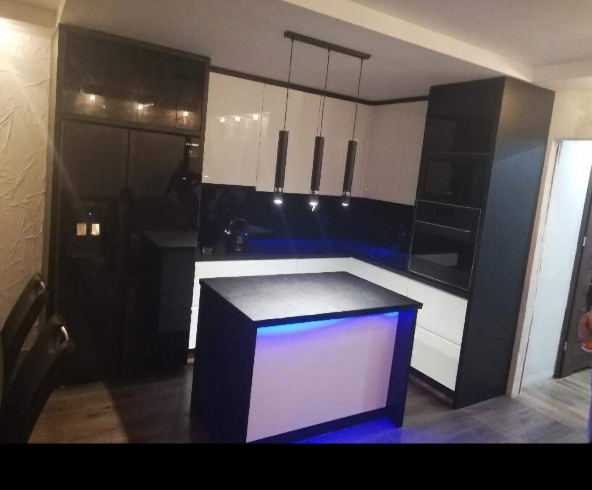 Nowoczesna kuchnia w czarno-białej kolorystyce z wyspą oświetloną niebieskim światłem LED, widoczne wiszące lampy i zabudowa kuchenna.