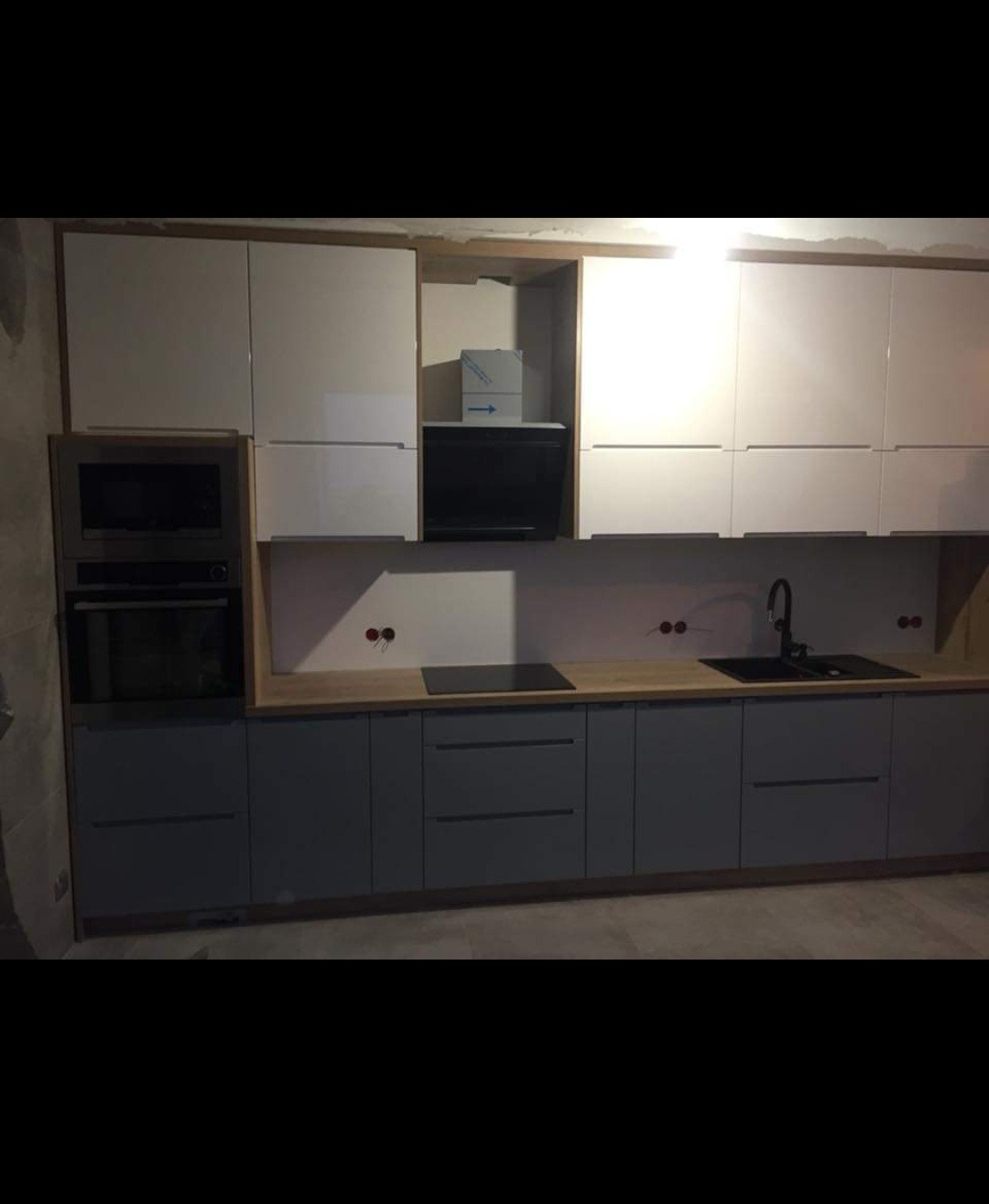 Nowoczesna, szara zabudowa kuchenna z białymi szafkami wiszącymi i drewnianym blatem, w trakcie montażu, widoczne niezakryte gniazdka elektryczne i kartonowe opakowanie po sprzęcie AGD.