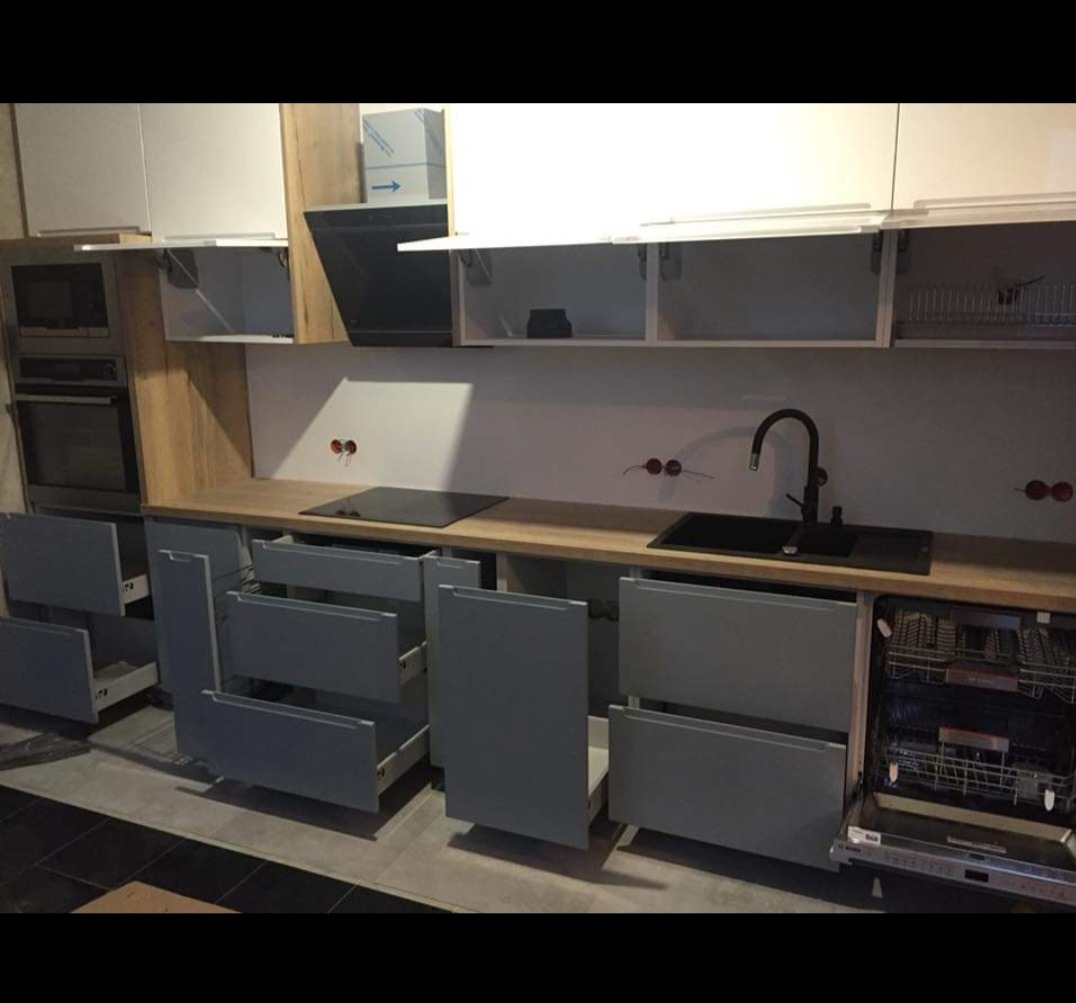 Nowoczesna kuchnia z szarymi szafkami i drewnianym blatem, otwarte szuflady i zmywarka, widoczne gniazdka elektryczne nad blatem.