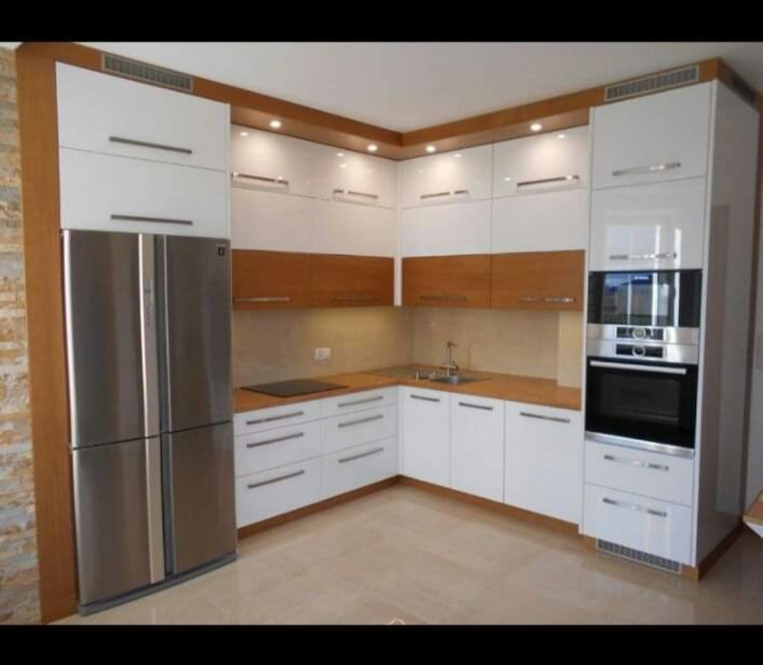 Nowoczesna kuchnia w kształcie litery L z białymi i drewnianymi frontami, oświetleniem LED podszafkowym i srebrną lodówką side-by-side, widok na narożnik i sprzęty AGD.