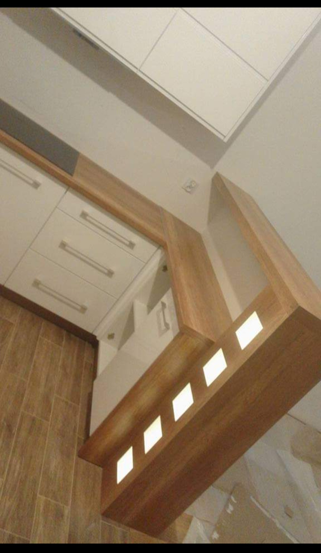 Nowoczesna kuchnia z białymi frontami szafek i drewnianym wykończeniem, widok z dołu ukazujący oświetlenie LED w drewnianej listwie pod szafkami.