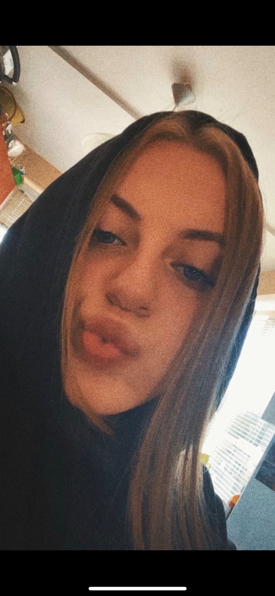 Portret młodej kobiety z blond pasemkami, ubrana w czarną bluzę z kapturem, robiąca selfie z lekkim uśmiechem.