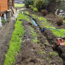 Wykopany rów w trawie z niebieskimi rurami, widoczne narzędzie do wykopów, dom z bali w tle. Prace ziemne związane z instalacją podziemną w ogrodzie.