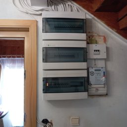 Mateusz Fryzowicz - Rozdzielnia elektryczna z licznikiem na ścianie, widoczne kable prowadzone wzdłuż sufitu, fragment futryny drzwi. Widoczny włącznik światła i otwór na gniazdko.