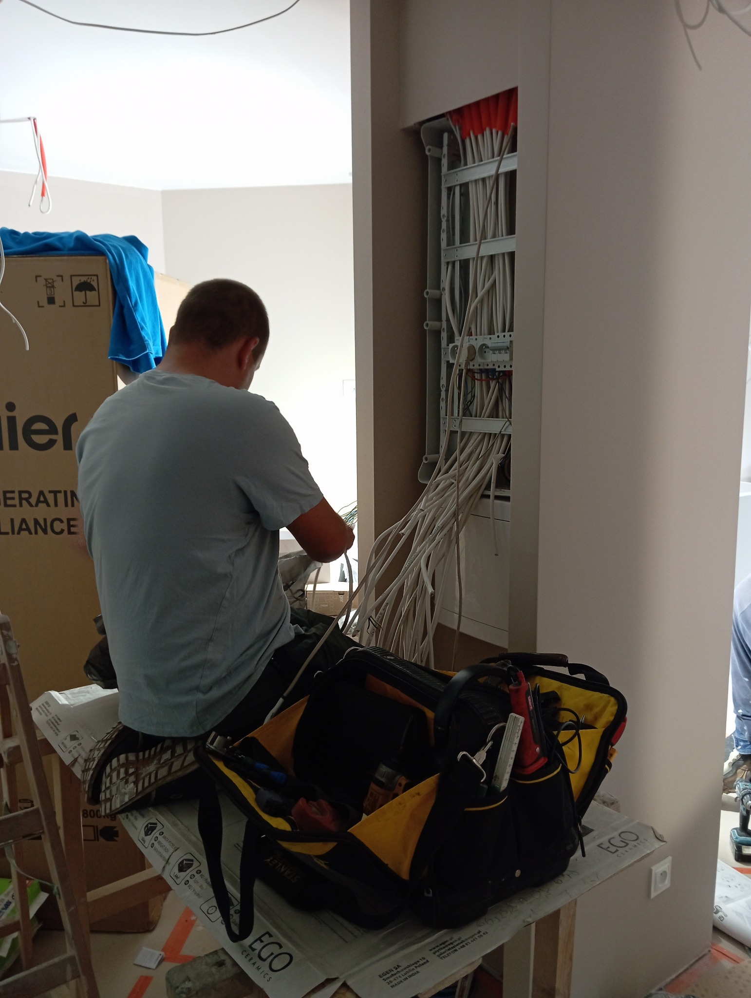 Elektryk w trakcie montażu instalacji elektrycznej w nowym budynku. Widoczne kable, skrzynka rozdzielcza i torba z narzędziami.