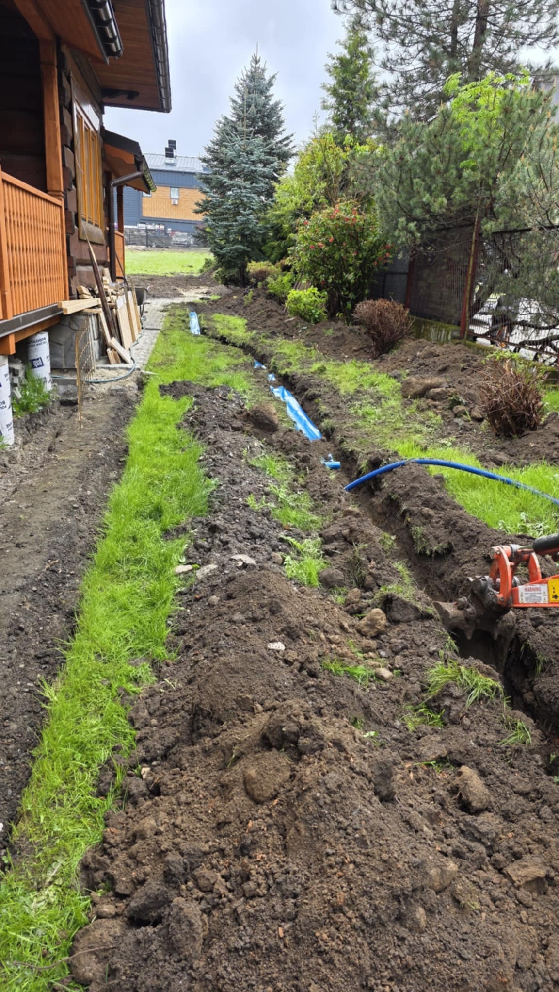Wykopany rów w trawie z niebieskimi rurami, widoczne narzędzie do wykopów, dom z bali w tle. Prace ziemne związane z instalacją podziemną w ogrodzie.