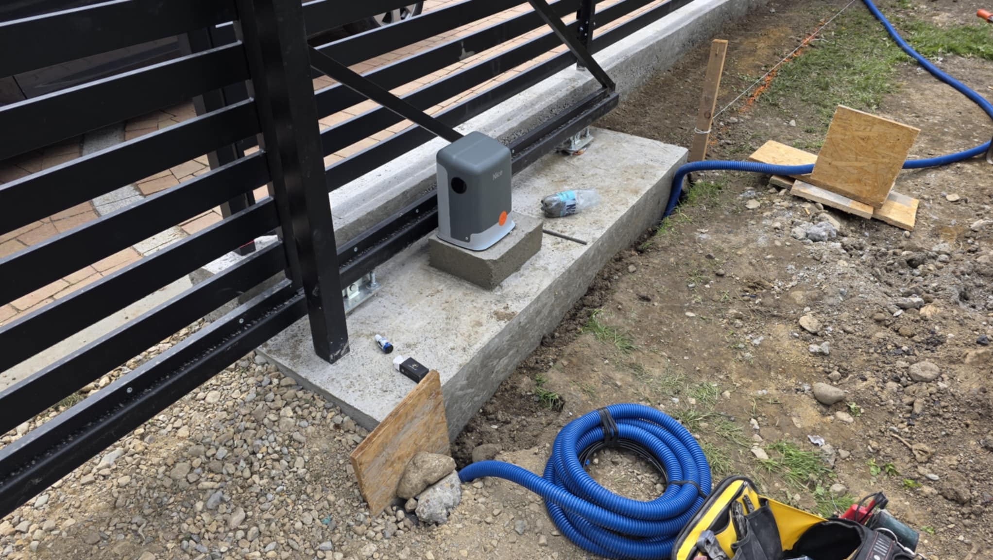 Montaż automatyki do bramy przesuwnej: szary napęd NICE na betonowym fundamencie, widoczne narzędzia i niebieski przewód. Elementy bramy w tle.