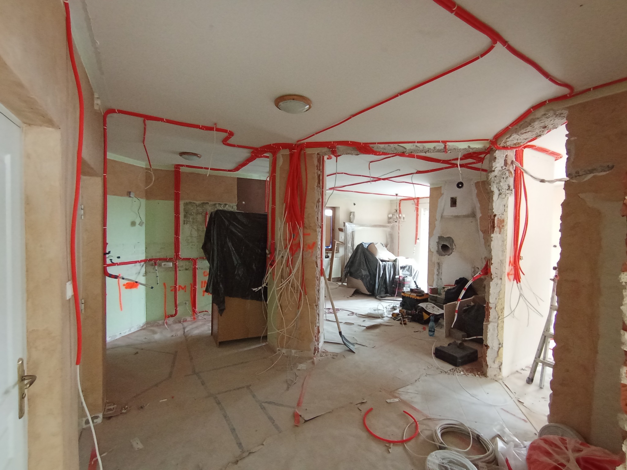 Rozprowadzenie instalacji elektrycznej w rurkach ochronnych, czerwone peszle na ścianach i suficie w surowym wnętrzu w trakcie remontu. Widoczne kable i narzędzia.