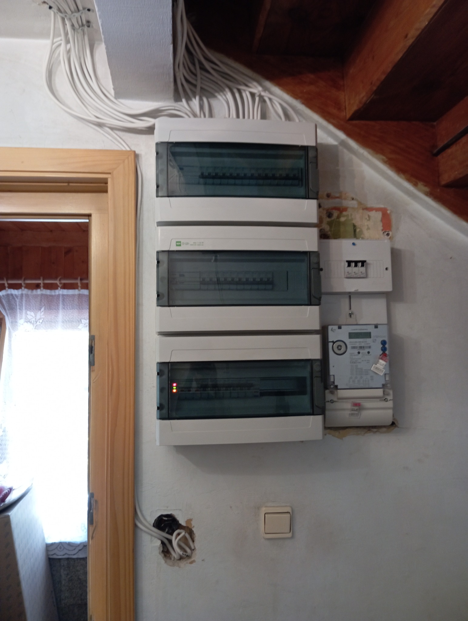 Rozdzielnia elektryczna z licznikiem na ścianie, widoczne kable prowadzone wzdłuż sufitu, fragment futryny drzwi. Widoczny włącznik światła i otwór na gniazdko.