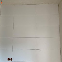 Konrad Sobol - Ściana wyłożona białymi, prostokątnymi płytkami ceramicznymi z widocznymi fugami, otwory pod gniazdka elektryczne i wystające kable z pomarańczowymi złączkami w górnym rogu.