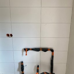 Konrad Sobol - Świeżo ułożone, białe płytki ścienne z widocznymi otworami na instalacje elektryczne i rury zabezpieczone czarną folią i pomarańczową taśmą.