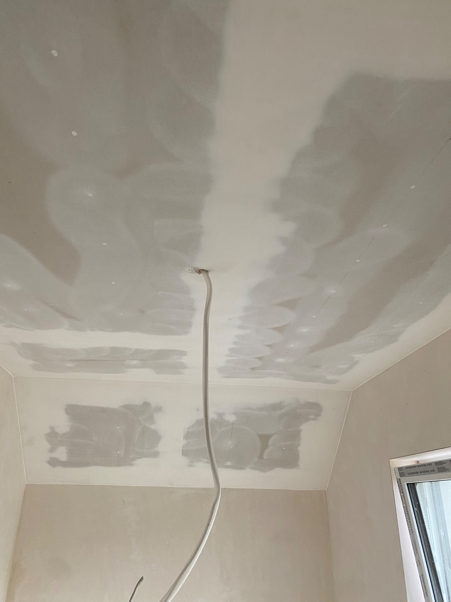 Sufit i górna część ściany po szpachlowaniu z widocznym przewodem elektrycznym wystającym z otworu w suficie i oknem w prawym dolnym rogu.