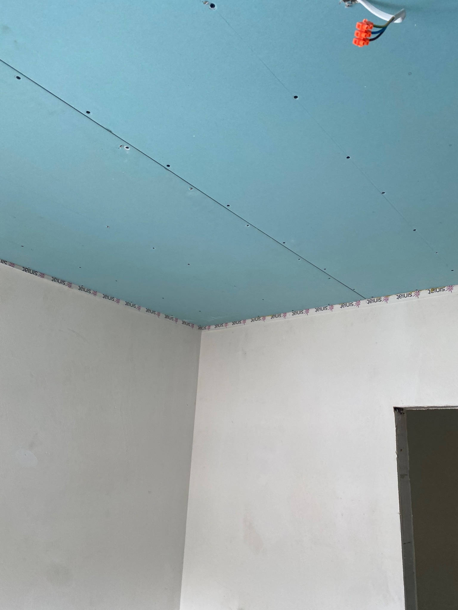 Niebieska płyta gipsowo-kartonowa zamontowana na suficie, z widocznymi śladami po wkrętach i taśmą marki Sinat na styku ze ścianą, z elementami instalacji elektrycznej wystającymi z sufitu.