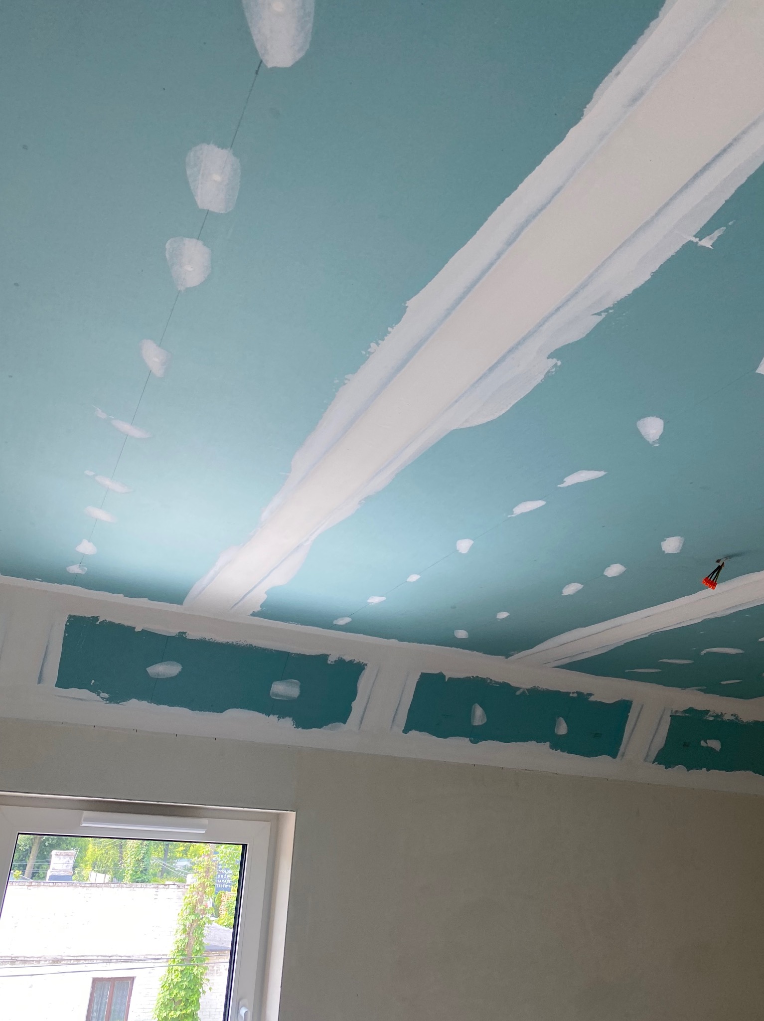 Niebieski sufit z zamontowanymi płytami, widoczne spoiny wypełnione białą masą szpachlową, przygotowany do dalszych prac wykończeniowych, okno w tle.