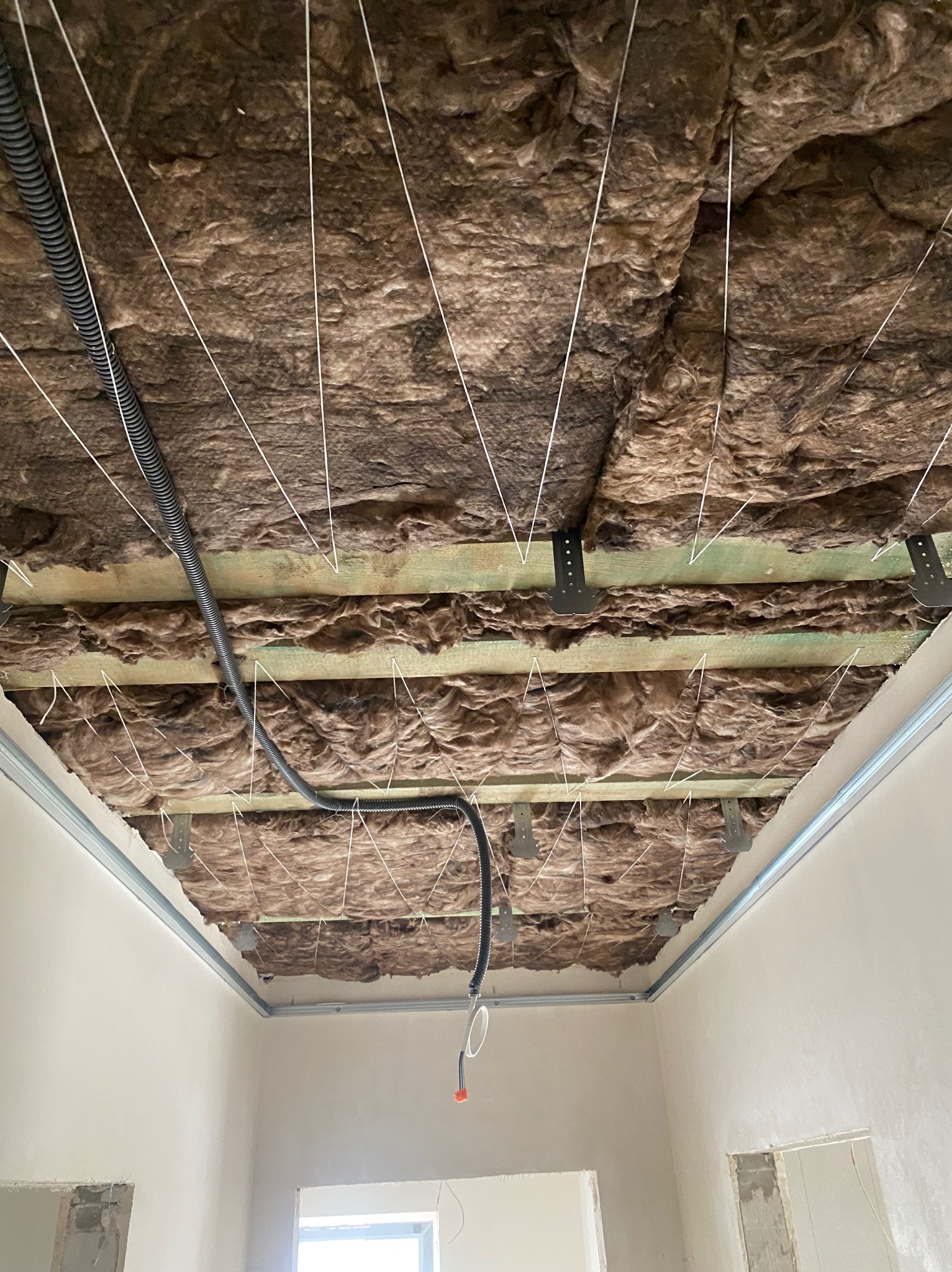 Poddasze w trakcie ocieplania wełną mineralną, widoczne drewniane belki, metalowe wieszaki, przewody elektryczne i sznurki podtrzymujące izolację, przygotowane pod montaż płyt.