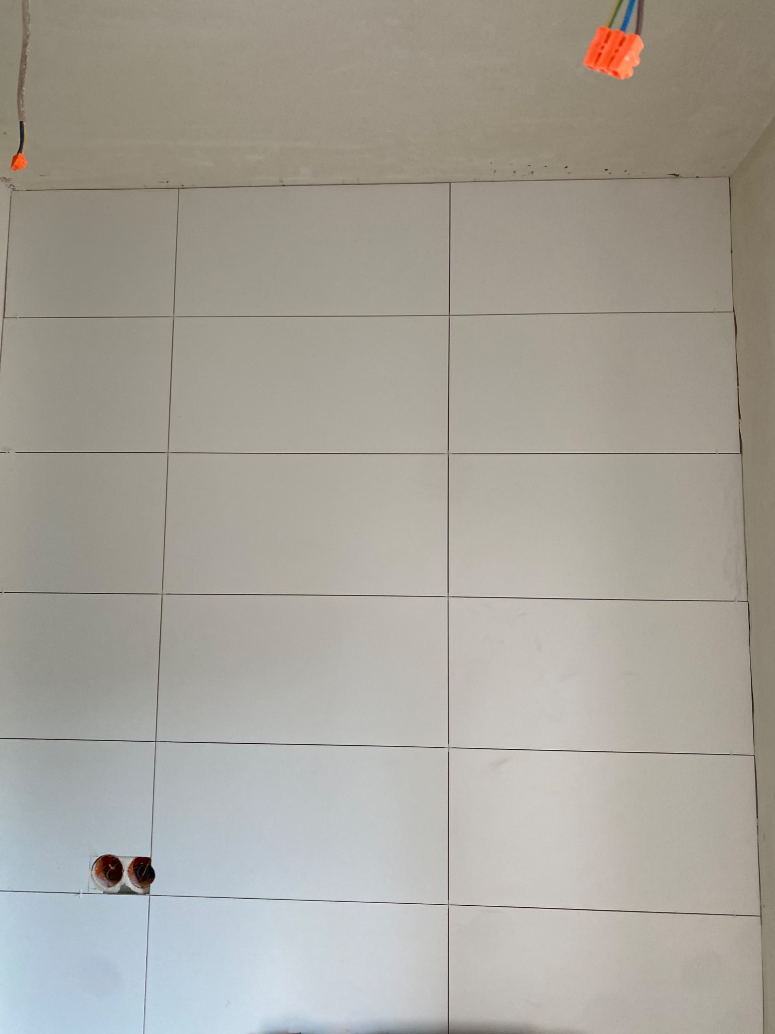 Ściana wyłożona białymi, prostokątnymi płytkami ceramicznymi z widocznymi fugami, otwory pod gniazdka elektryczne i wystające kable z pomarańczowymi złączkami w górnym rogu.
