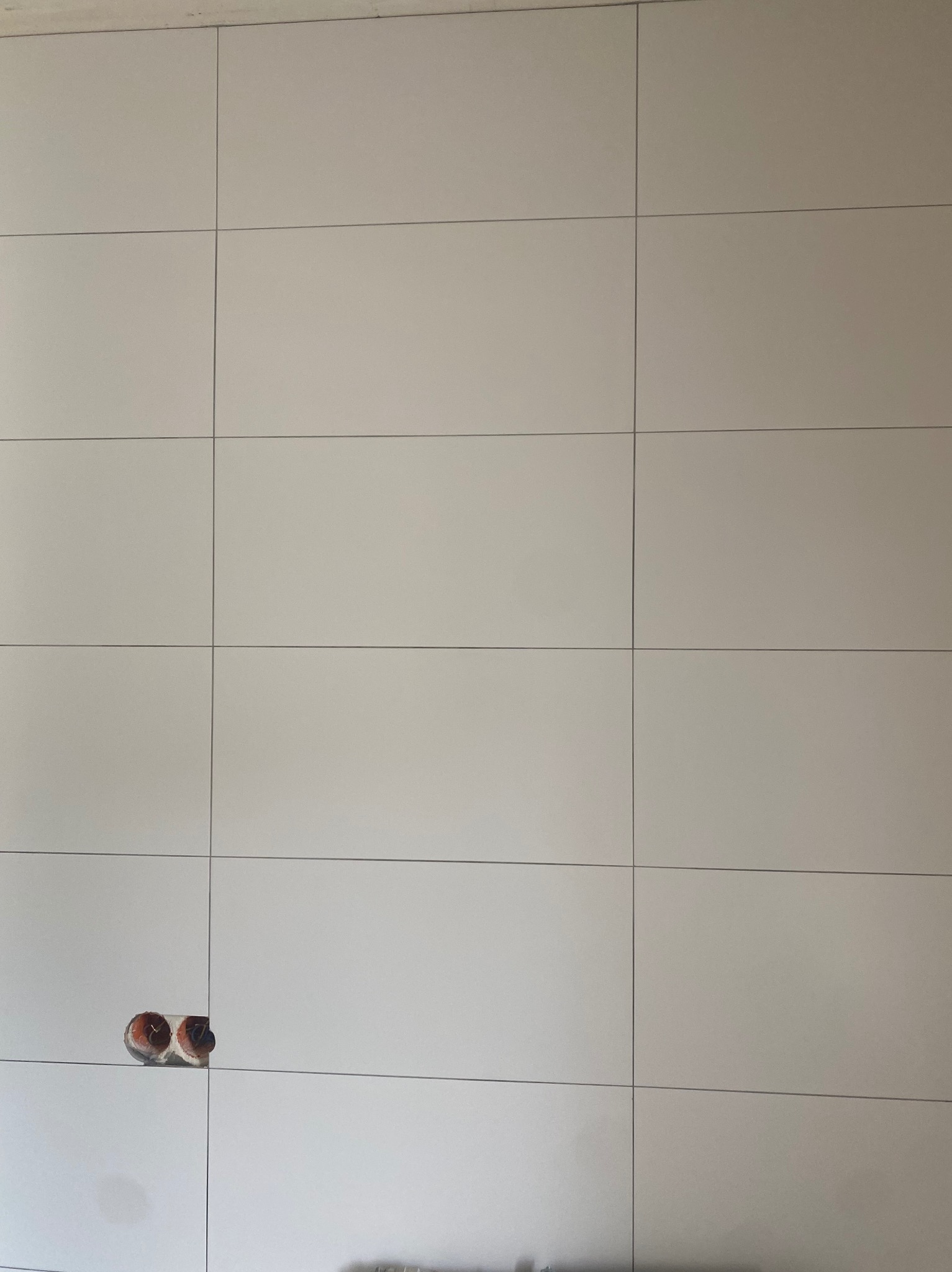 Ściana wykończona jasnymi, kwadratowymi płytami z widocznymi fugami i gniazdkami elektrycznymi bez osprzętu w dolnej części.