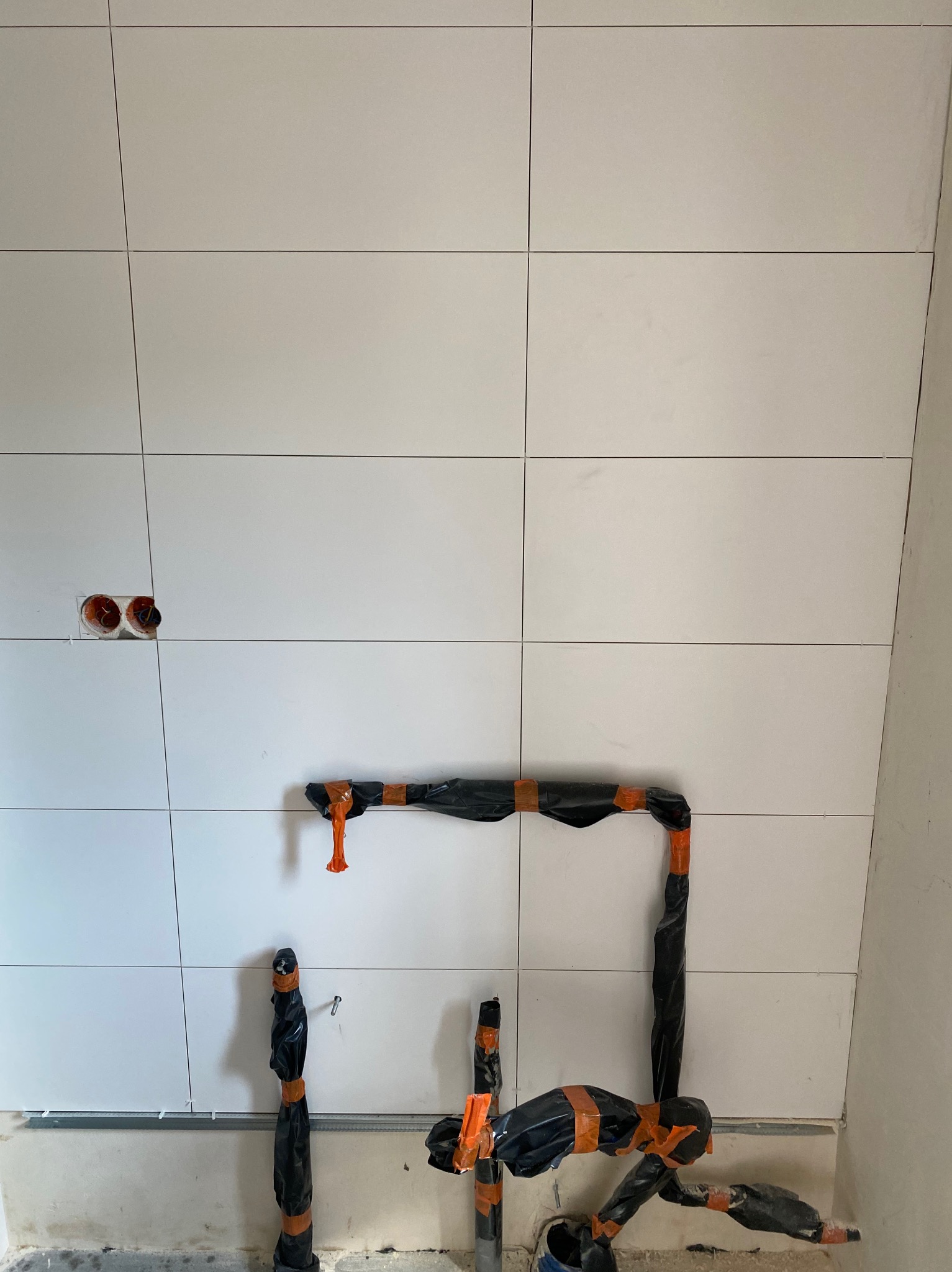 Świeżo ułożone, białe płytki ścienne z widocznymi otworami na instalacje elektryczne i rury zabezpieczone czarną folią i pomarańczową taśmą.