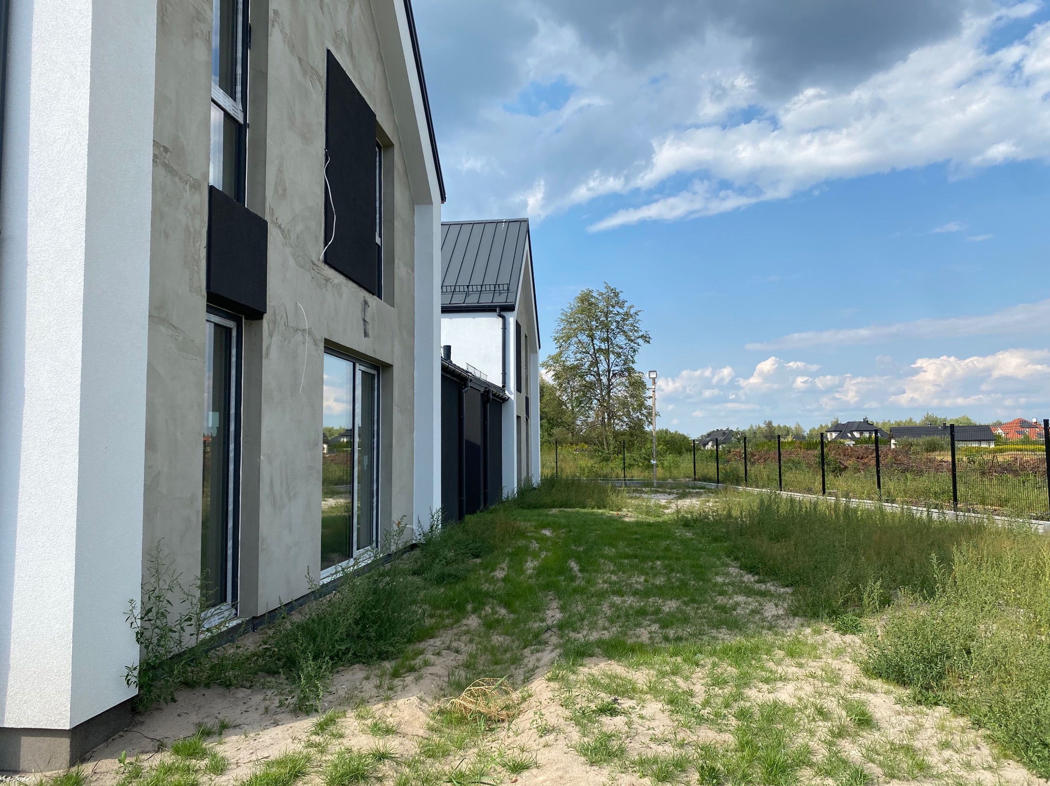 Nowoczesny dom w trakcie wykończenia elewacji, widoczne surowe ściany z oknami, czarne elementy dekoracyjne i nierówny teren wokół budynku w słoneczny dzień.