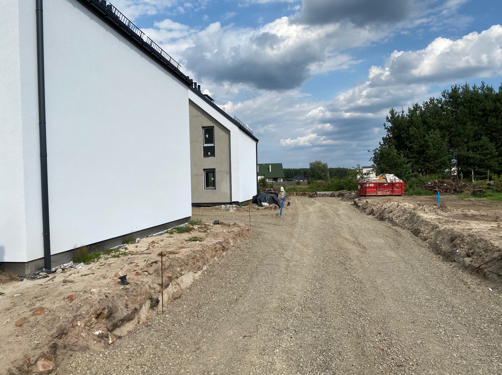 Nowoczesny dom jednorodzinny z białą elewacją, widok z poziomu gruntu na podjazd z utwardzonego kruszywa, w tle błękitne niebo z chmurami i fragment lasu.