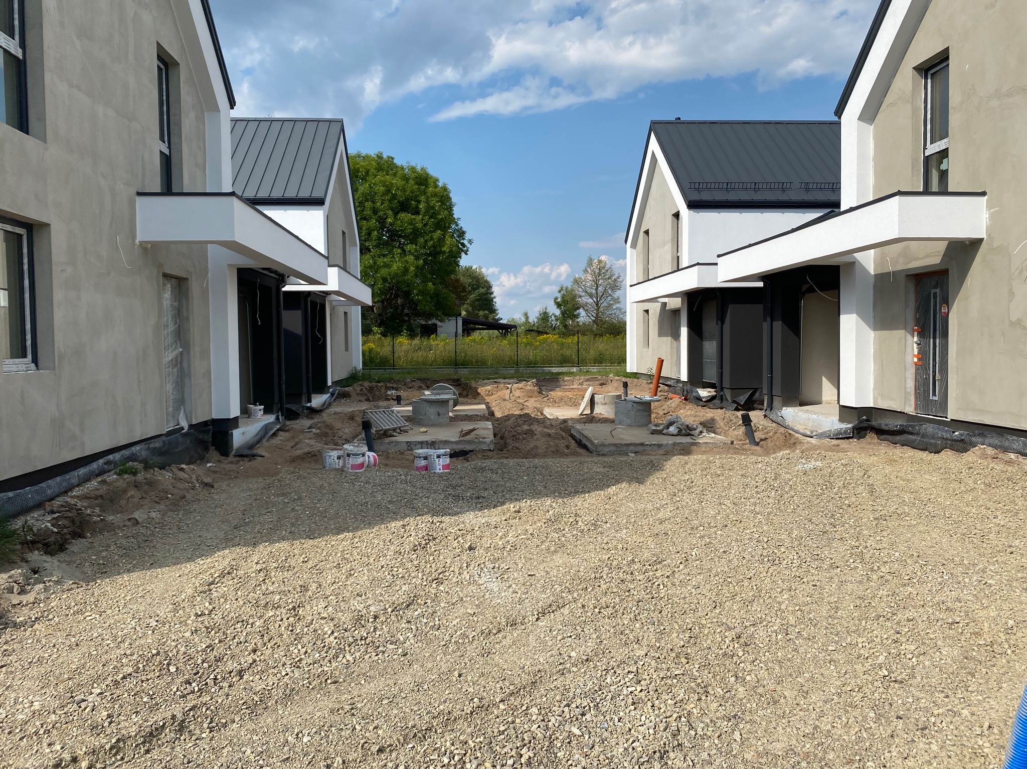 Nowe domy w trakcie budowy: surowe elewacje, szary dach, teren przygotowany pod wjazd, widoczne puszki farb i elementy instalacji.